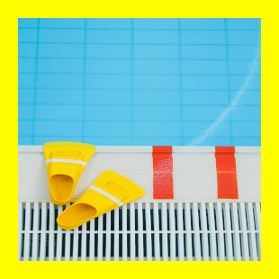 Schwimmbecken mit klarem, blauen Wasser von oben betrachtet, man sieht den Rand des Beckens und die Abdeckung des Wasserüberlaufs im unteren Bildteil, auf dem Beckenrand liegen gelbe Schwimmflossen und man sieht zwei orangene Streifen aufgeklebt
